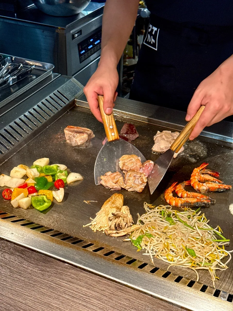 木の柄のヘラ二本で、肉とエビ、もやしを鉄板の上で同時に扱う料理人の両手。エプロンに2×2の字母マークが入っている