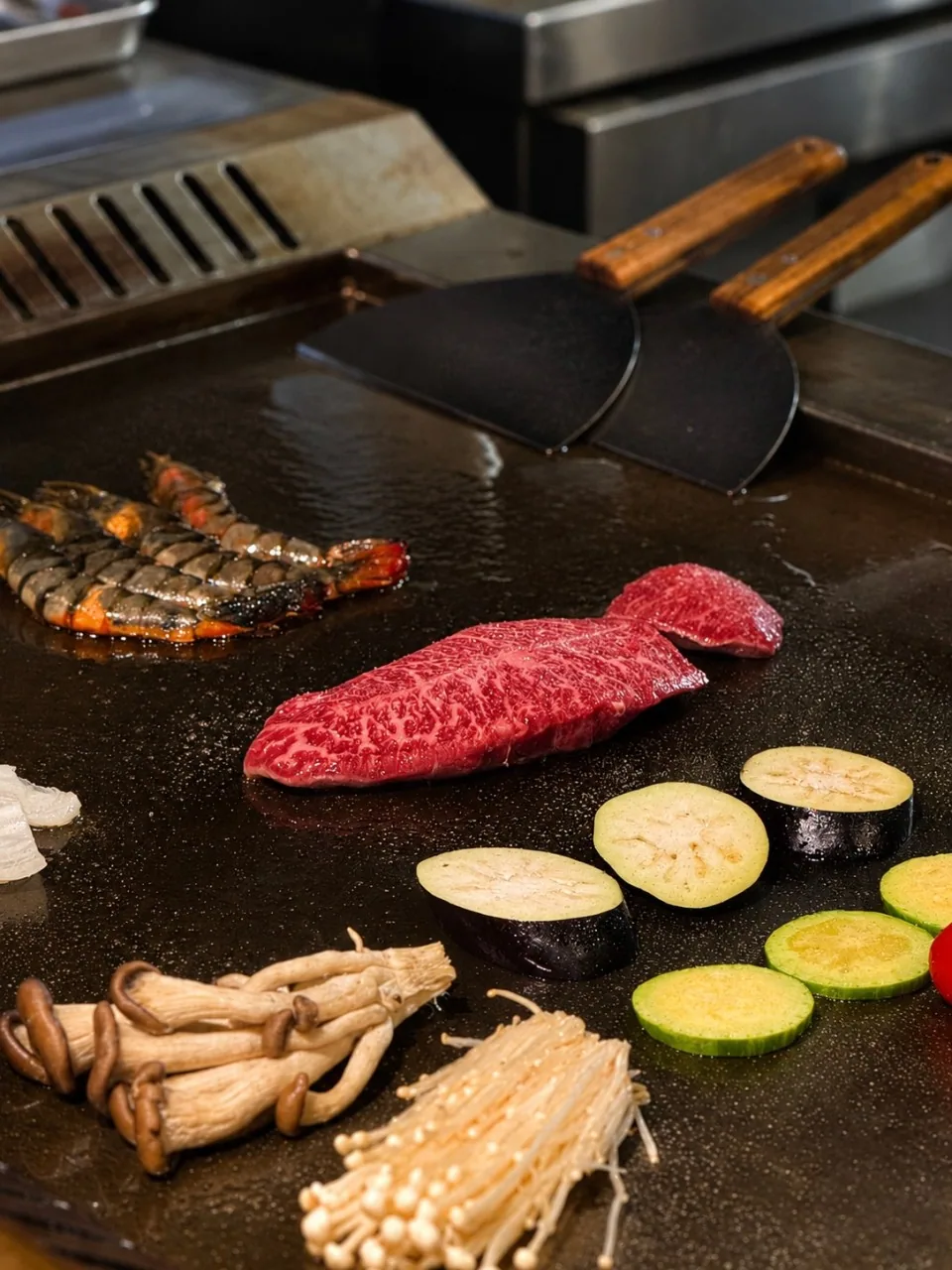 細かなサシの入った肉一枚と、大ぶりのエビ、ナス・ズッキーニ・きのこが並ぶ鉄板と、木の柄のヘラ二本