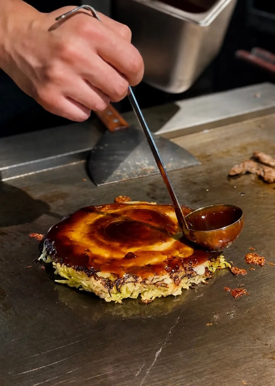 焼き上がったお好み焼きに、お玉を傾けて濃い茶色のソースをかける手
