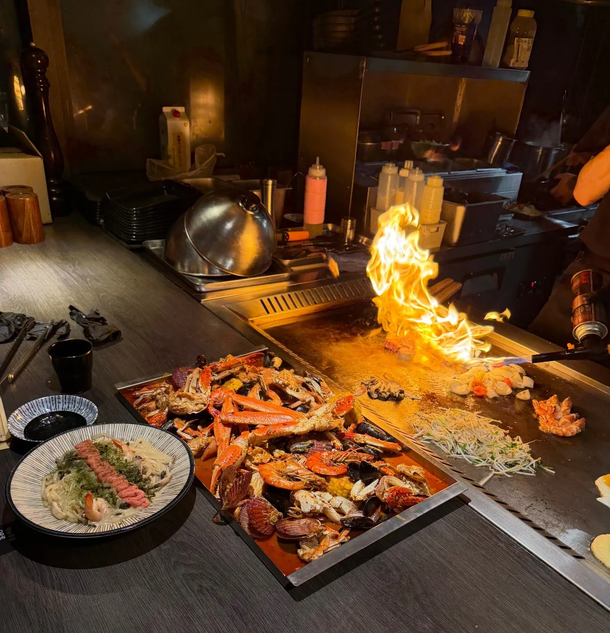 鉄板から立ち上がる炎の横に、カニの脚とムール貝・エビ・とうもろこしを盛ったトレー